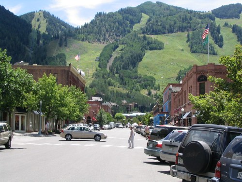 A scenic street view with shops and parked cars, framed by green mountains and blue sky in a small town.