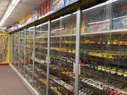 A display of colorful toy cars and models in glass cases, with shelves of boxes above in a well-lit store.
