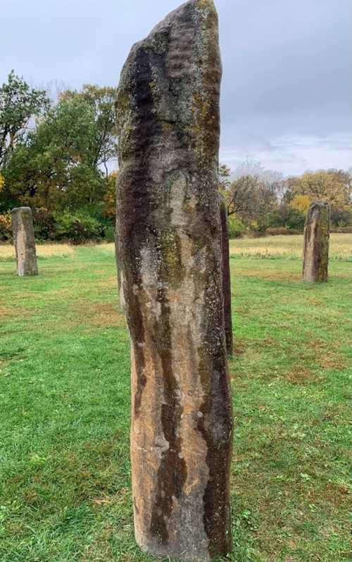 A tall, weathered stone standing among shorter stones in a grassy field, with trees in the background under a cloudy sky.