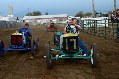 Two vintage race cars compete on a dirt track, with one driver focused and another in the background.