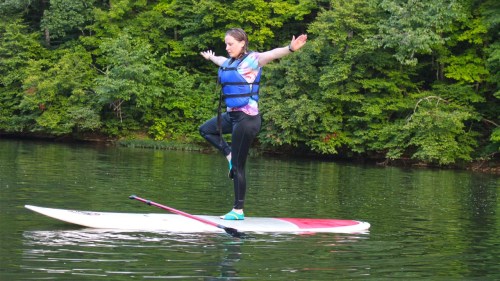 A person balances on a paddleboard in a lake, surrounded by lush green trees, wearing a life jacket.