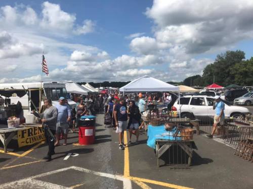 A bustling outdoor market with people browsing stalls, tents, and vehicles under a partly cloudy sky.
