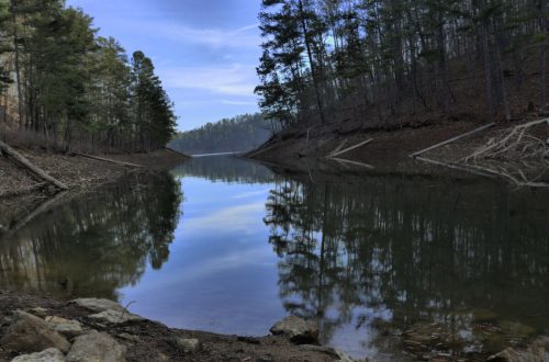 A calm river reflects trees and a blue sky, surrounded by a wooded landscape and rocky banks.
