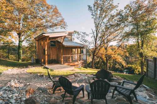 A cozy wooden cabin surrounded by autumn trees, with a stone patio and chairs in the foreground.