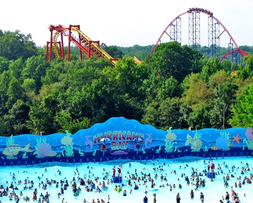 A busy water park with a wave pool and a roller coaster in the background, surrounded by lush greenery.