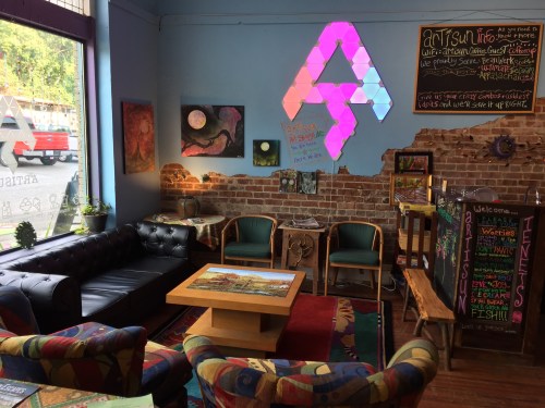 Cozy café interior with colorful seating, brick walls, art displays, and a vibrant light installation.