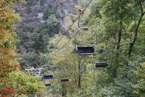 A ski lift with empty chairs suspended above a forested area with autumn foliage.