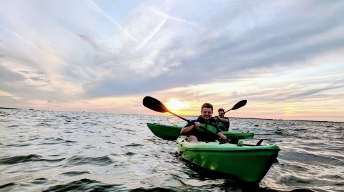 Two people in green kayaks paddle on a calm sea during sunset, with colorful clouds in the sky.