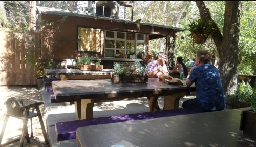 Outdoor dining area with wooden tables, people eating, and a rustic building surrounded by trees.