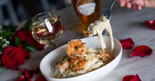 A plate of creamy shrimp pasta with a fork, accompanied by a glass of white wine and a beer, surrounded by rose petals.
