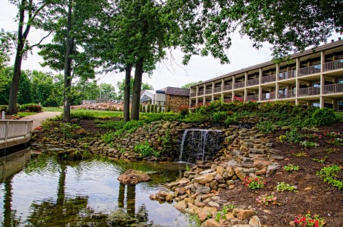 A serene landscape featuring a small waterfall, rocks, and a building surrounded by trees and colorful flowers.
