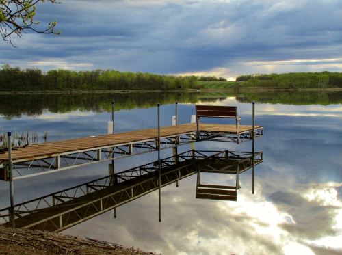 A serene lake scene with a wooden dock and bench, reflecting clouds and trees in calm water under a cloudy sky.