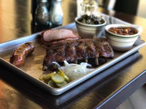 A metal tray with barbecue ribs, sausage, pickles, onions, greens, and baked beans on a wooden table.