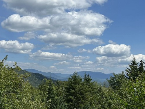 A scenic view of mountains under a blue sky with fluffy clouds, surrounded by lush green trees.