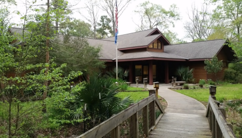 A wooden building surrounded by greenery, with a pathway leading to the entrance and an American flag in front.