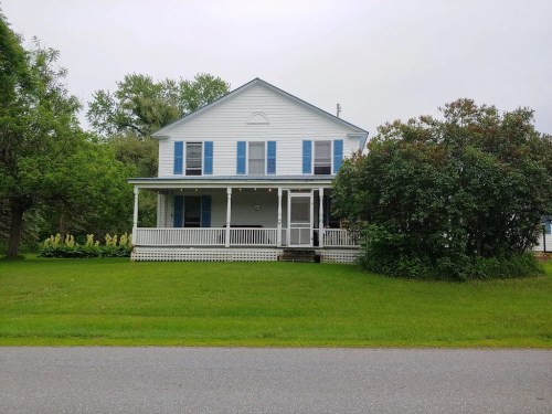 A two-story white house with blue shutters, a front porch, and a green lawn, surrounded by trees.