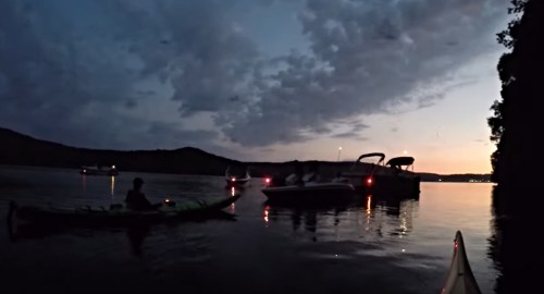 A kayaker paddles near boats on a calm lake at dusk, with clouds and a fading sunset in the background.