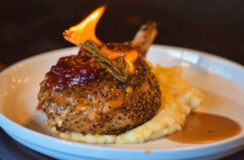 A seared pork chop topped with cranberry sauce and rosemary, served on creamy mashed potatoes, with flames flickering.