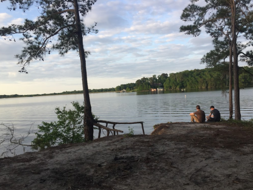 Two people sit by a calm lake, surrounded by trees, with a cloudy sky reflecting on the water's surface.
