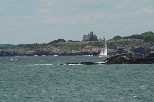 A sailboat glides across the water near a rocky shore, with a distant castle on a green hill under a cloudy sky.