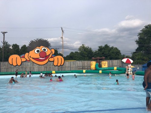 A colorful pool scene with children swimming, featuring a large cartoon character on the wall and a water slide.