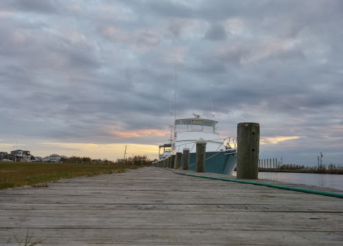 A wooden dock leads to a boat moored at sunset, with cloudy skies and grassy areas in the background.