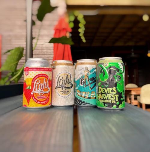 Four colorful beer cans arranged on a table, featuring various designs and labels, with a blurred outdoor background.