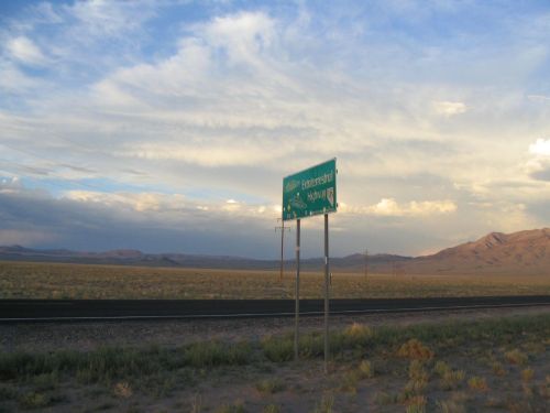 A green highway sign stands beside a road in a vast, open landscape under a cloudy sky. Mountains are visible in the distance.