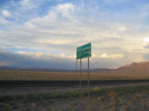 A green highway sign stands beside a road in a vast, open landscape under a cloudy sky. Mountains are visible in the distance.