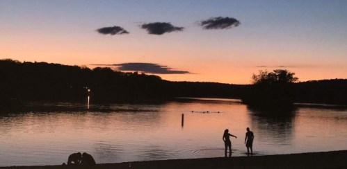 Silhouettes of two people by a calm lake at sunset, with soft clouds and a serene landscape in the background.