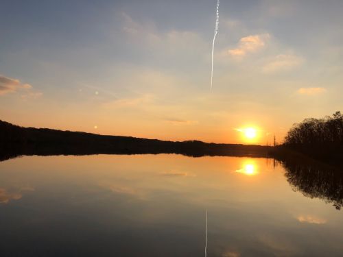 A serene sunset over a calm lake, reflecting the sky and trees, with a faint contrail above.