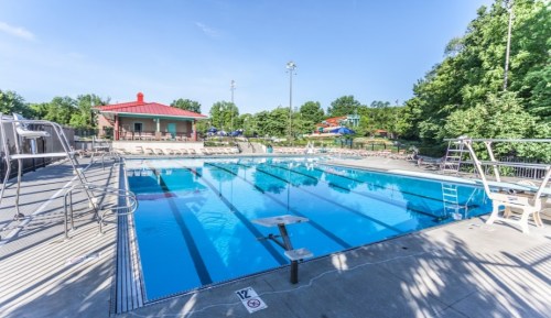 A clear blue swimming pool surrounded by lounge chairs and greenery, with a red-roofed building in the background.