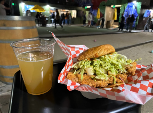 A fried chicken sandwich with lettuce on a checkered paper, alongside a cup of light beer, in a lively outdoor setting.