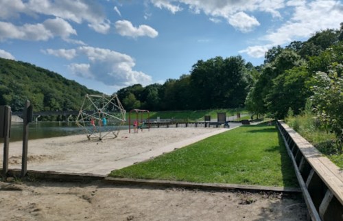 A sandy beach area by a lake, with playground equipment and green trees under a partly cloudy sky.