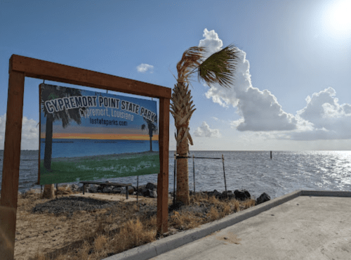 Sign for Cypremort Point State Park in Louisiana, with palm trees and a view of the water under a cloudy sky.