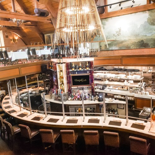 A spacious restaurant interior featuring a circular bar, elegant decor, and a large chandelier overhead.