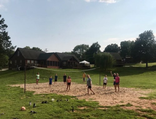 A group of people playing beach volleyball on a sandy court with cabins and trees in the background.