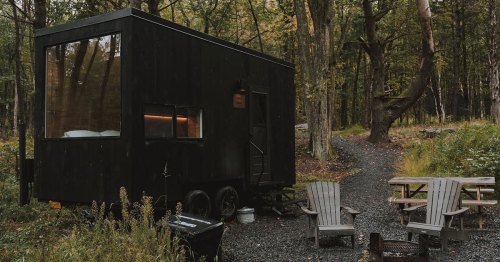 A modern black tiny house on wheels surrounded by trees, with a gravel path and outdoor seating area nearby.