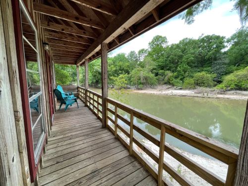 A wooden porch overlooking a calm river, surrounded by lush greenery and trees. Two blue chairs are visible.