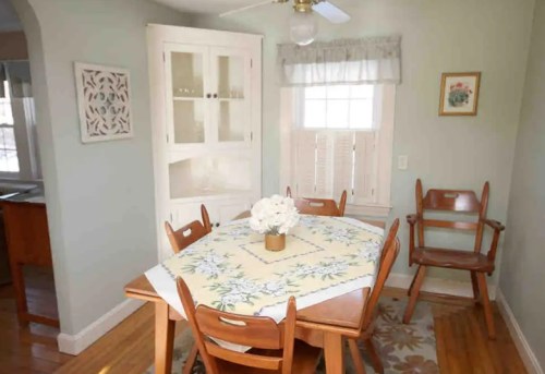 Cozy dining area with a wooden table, chairs, and a floral tablecloth, featuring a window and light-colored walls.