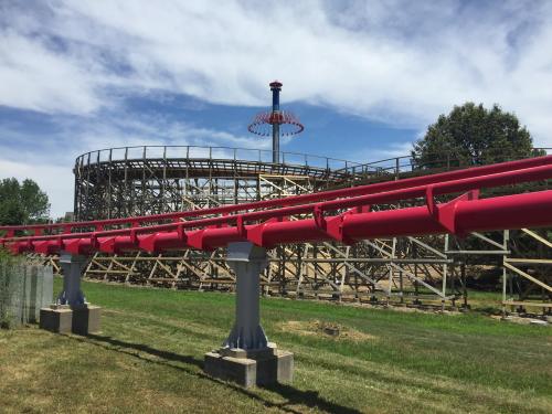 A red roller coaster track curves in front of a wooden coaster under a blue sky with scattered clouds.