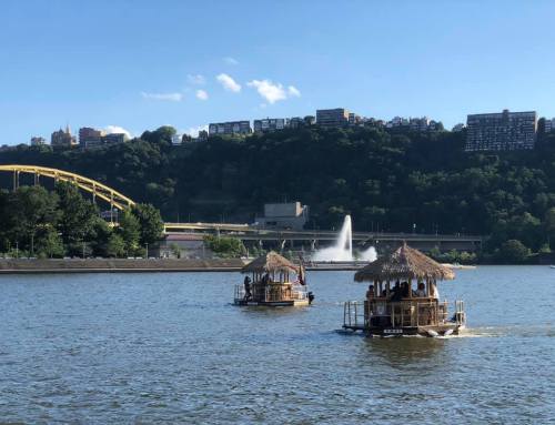 Two tiki-style boats float on a river, with a fountain and city skyline in the background under a clear blue sky.