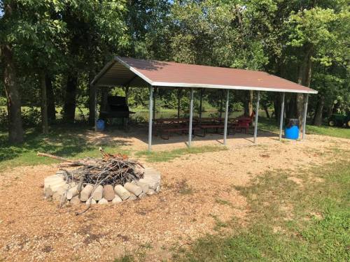 A covered picnic area with a fire pit and seating, surrounded by trees and grass.