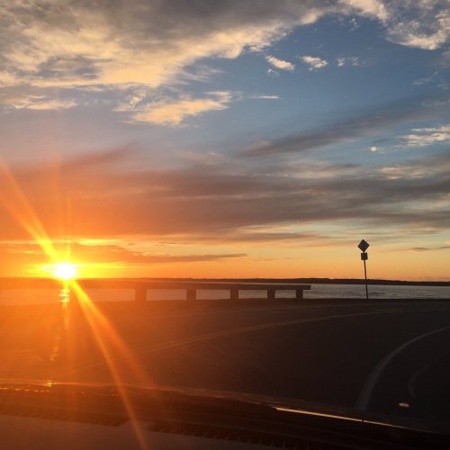 A vibrant sunset over a calm body of water, viewed from a winding road with a sign in the distance.