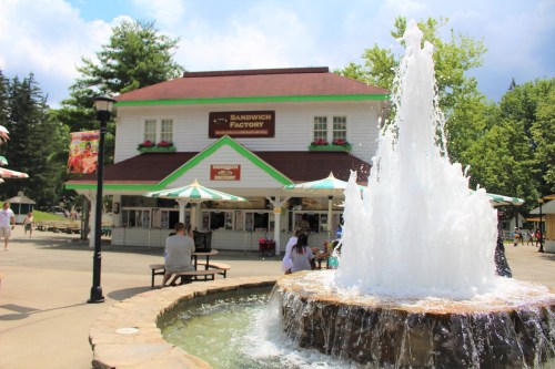 A fountain in front of a building labeled "Sandwich Factory," with outdoor seating and colorful umbrellas.