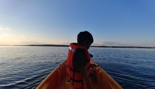 A child in an orange life jacket sits in a kayak on calm water, with a clear blue sky and distant shoreline.