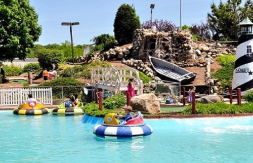 Colorful bumper boats in a water attraction, surrounded by greenery and a mini-golf course with a lighthouse.