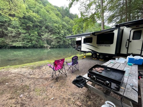 A campsite by a serene lake with a camper, picnic table, and grilling burgers on a flat top grill.