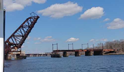 A rusted drawbridge partially raised over a river, with blue skies and fluffy clouds in the background.