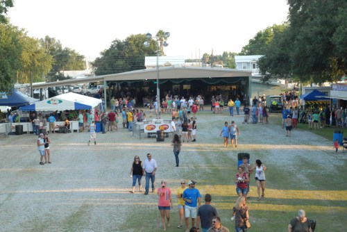 A lively outdoor festival scene with people walking, food stalls, and a large gathering under a pavilion.